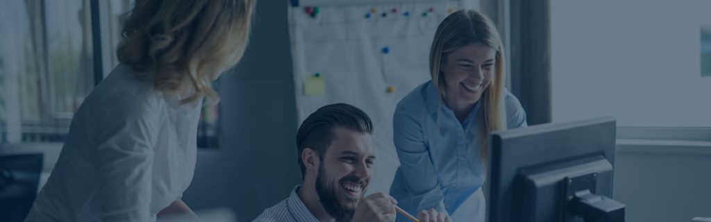An office environment with a man sat at a desk looking at a monitor. He is smiling. At each side of him are two blonde women standing up in professional clothing, also looking at the screen and smiling.