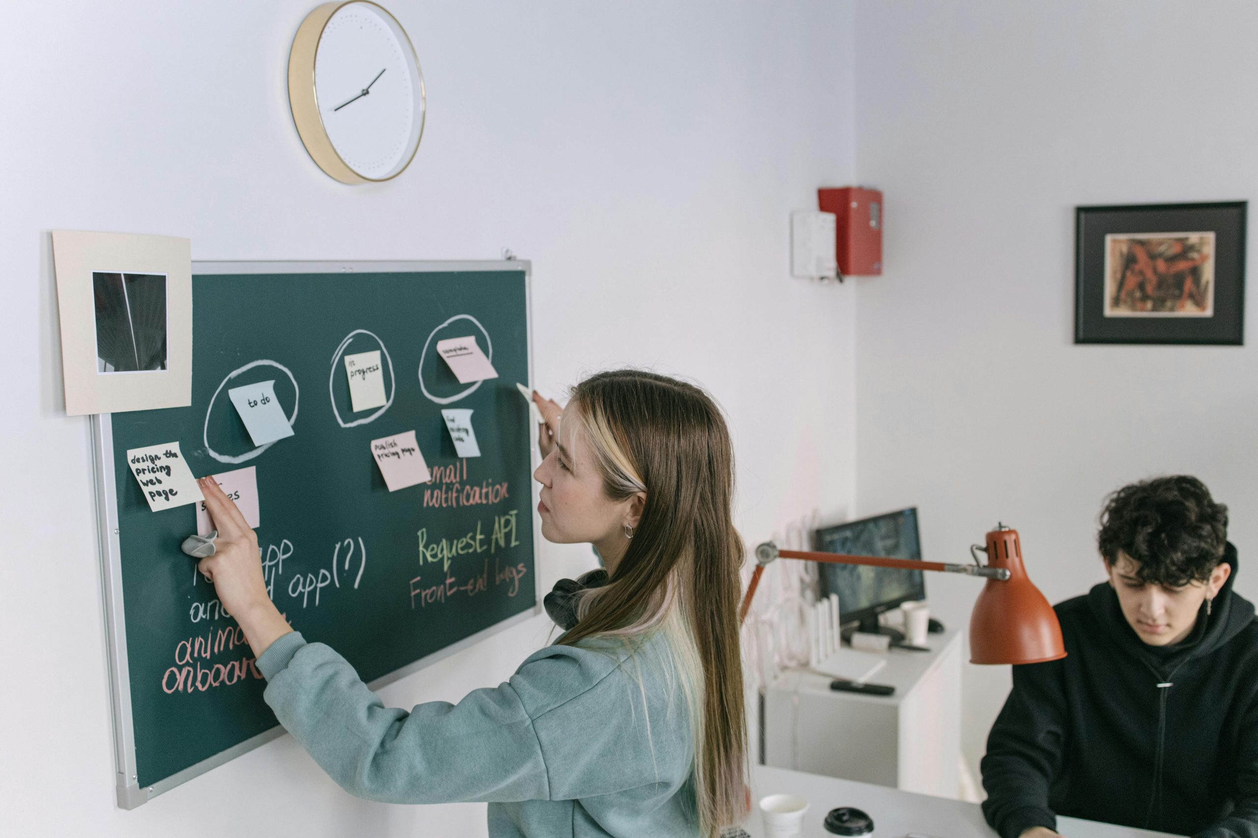 An office setting. Two young entrepreneurs are writing on a board which has post-it notes stuck to it. They look like they need administrative support or a virtual assistant for their small business.